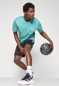Camisa de manga corta color teal con logo verde, pantalones cortos negros con rayas blancas, balón de baloncesto negro y zapatillas deportivas negro/blanco con suelas prominentes.