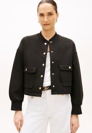 Frau mit zurückgebundenem Haar, die eine schwarze, geknöpfte Jacke mit Pattentaschen, weiße Hose und einen Gürtel mit goldenen Initialen trägt.