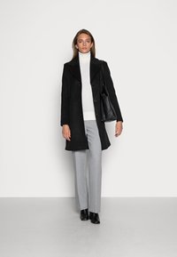 Melnas vilnas mētelis ar lielu apkakli un pogu aizdari, uzvilkts virs baltas turtlenecks, pelēkiem piegrieztiem biksēm un melniem potīšu zābakiem, aksesuāri ar melnu somu.