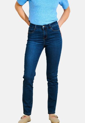 Jean skinny en denim extensible bleu foncé avec cinq poches, passants de ceinture et coutures contrastées. Porté avec un haut bleu clair et des chaussures beiges.