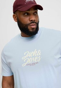 Lyseblå t-shirt med korte ærmer, der har "JACK & JONES" logo i hvid og "WORLDWIDE STUDIO" i rød, båret med en burgunder kasket.