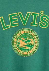 Grønn bomull t-skjorte med fet gul "LEVI'S" tekst og sirkelformet logo med fjell og sol, omkranset av en bølget kant.