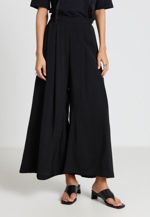 Donna che indossa pantaloni neri a vita alta e gamba larga, top nero a maniche corte, sandali neri con tacco e anelli d'argento su entrambe le mani.