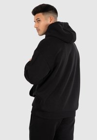Haut à capuche en coton noir avec une coupe décontractée, des manches surdimensionnées, des poignets côtelés et un capuchon avec cordon de serrage, vu de côté.