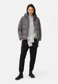 Grå dunjakke med hætte, lynlås og elastiske manchetter. Båret over en lys grå sweater og sorte tapered bukser, suppleret med sorte støvler.
