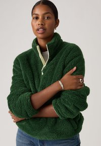 Γυναίκα με σκούρα μαλλιά φοράει πράσινο σακάκι από φλις με φερμουάρ στο μισό, μπλε τζιν, ασημένιο βραχιόλι και δαχτυλίδι, στέκεται μπροστά σε απλό φόντο.