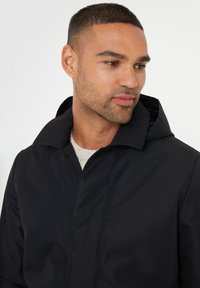 Zwarte hoodie met een gladde textuur, met een eenvoudig kraagontwerp en een sluiting aan de voorkant. Draagt over een lichtgekleurde trui.