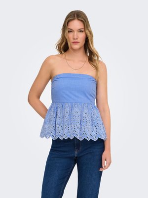 Femme portant un top bleu bustier avec un ourlet en dentelle ajourée brodée et un jean bleu foncé, debout devant un fond uni.
