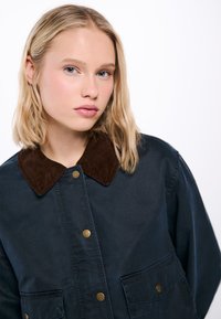 Mörkblå jacka med brun kordflodorkrage, metallknappar och två framfickor. Tyget verkar slätt, med en avslappnad design.