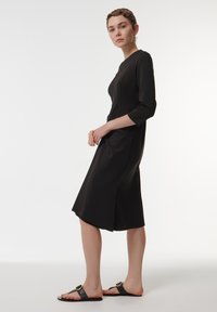 Vestido negro con mangas tres cuartos, corsé ajustado y falda acampanada. Tela suave con un acabado mate. Llevado con sandalias negras planas.