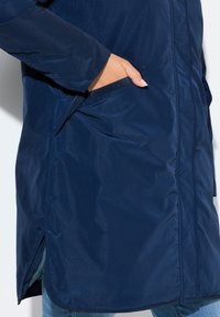 Laivastonsininen, vettähylkivä pitkä takki, jossa on sileä pinta, sivutaskut ja helmassa hienovaraiset halkiot liikkuvuuden helpottamiseksi.