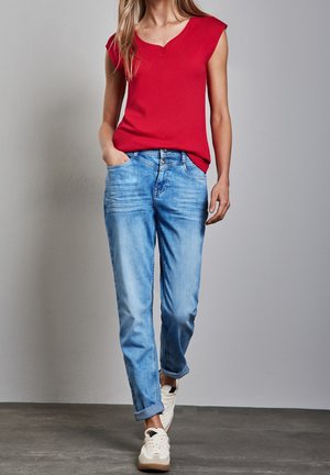 Frau trägt ein rotes kurzärmeliges Oberteil, hellblaue hochgekrempelte Jeans und weiße Turnschuhe, steht mit einer Hand in der Tasche vor einer einfarbigen Wand.