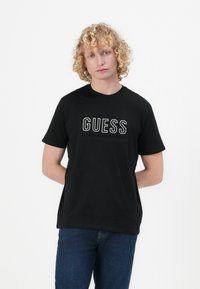 Svart bomull T-skjorte med en stor hvit "GUESS" logo på forsiden, rund hals og korte ermer. Bært med blå jeans.
