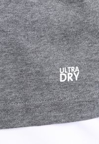 Šedá pletenina s bílým logem "ULTRA DRY" potisknutým blízko lemu, ukazující texturu a detaily šití.