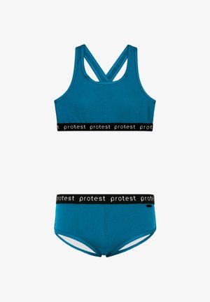 Tirkizni sportski grudnjak i odgovarajuće gaćice od teksturiranog materijala. Crni pojas sadrži ponovljeno "protest" brendiranje. Dizajn s križem na leđima za podršku.