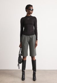 Black long-sleeve top with ruched detailing, green tailored shorts, knee-high black boots, and a small black bag. Accent sunglasses and earrings.