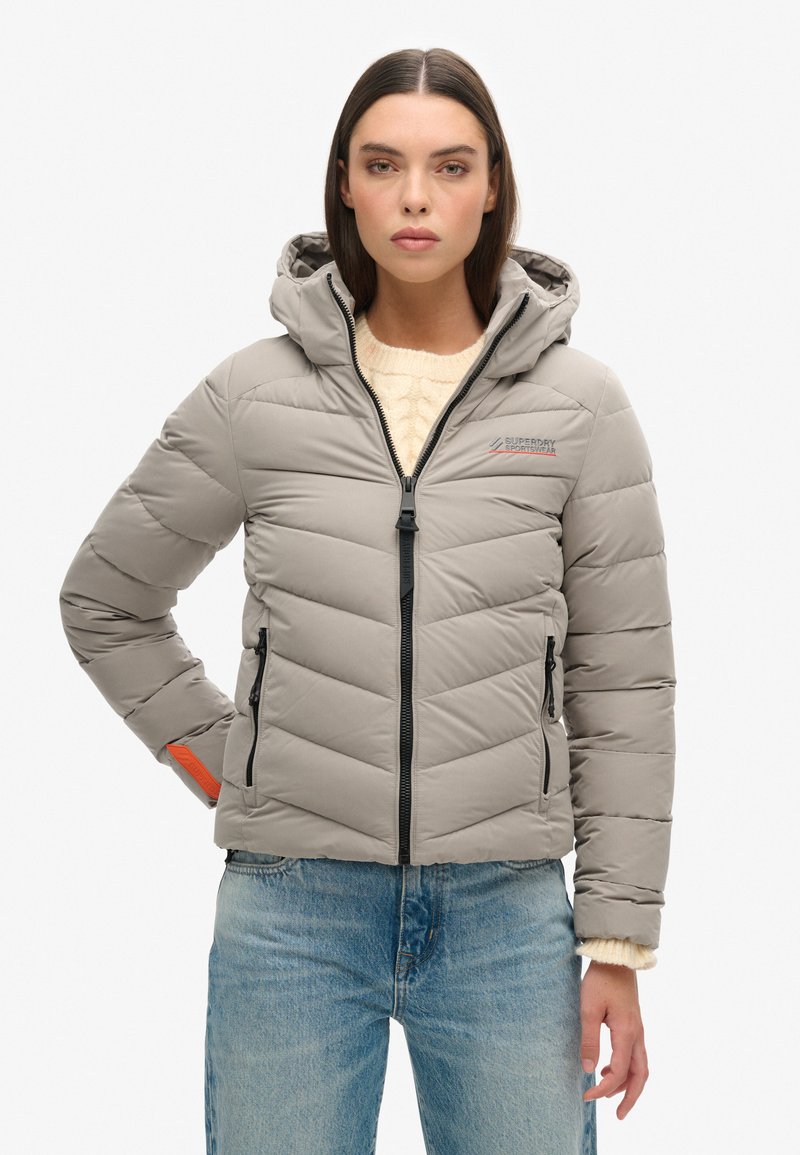 Doudoune grise avec fermeture éclair à l'avant, capuche et coutures en chevron. Elle possède deux poches zippées et un accent orange distinctif sur le poignet.