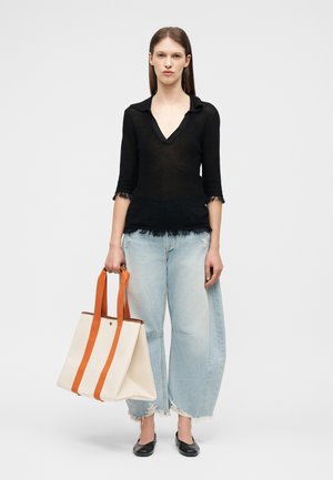 Mladá žena v černém topu s třásněmi a širokých světle modrých džínách, držící velkou béžovou nákupní tašku s oranžovými popruhy, stojící na jednobarevném pozadí.