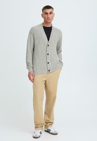 Svetle šedý žebrovaný cardigan s výstřihem do V, zapínáním na knoflíky a dlouhými rukávy, v kombinaci s béžovými kalhotami a bílými teniskami. Jednoduchý, neformální design.
