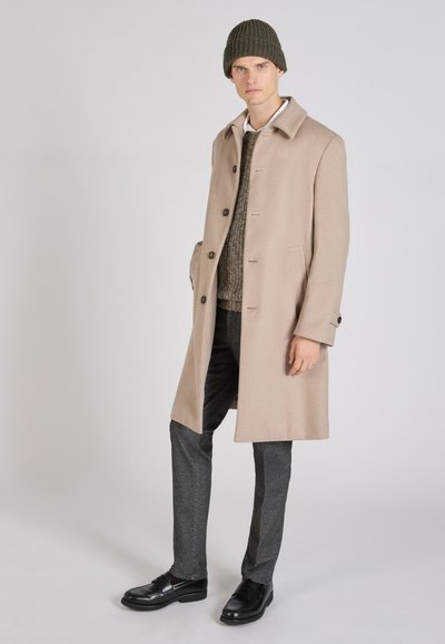 Beige wollen jas met een getailleerde pasvorm, voorzien van een gelapte kraag, vijf knopen en zijzakken. Gedragen over een groene gebreide trui en grijze broeken.