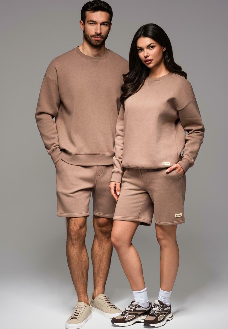 Dwie modelki noszą dopasowane beżowe bluzy i szorty z miękkiego materiału. Bluzy mają prążkowane mankiety, a oba outfity zawierają kieszenie.