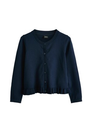 Junges Mädchen steht zwischen zwei Holzstühlen, trägt eine marineblaue Strickjacke, ein weißes Hemd mit Kragen, einen Faltenrock, weiße Socken und schwarze Schuhe.