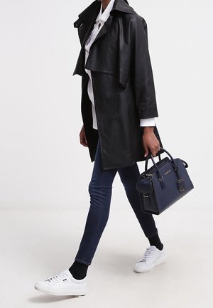 Personne marchant, portant un trench-coat en cuir noir, une chemise blanche, un jean skinny foncé, des baskets blanches, et portant un sac à main bleu marine.