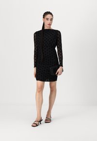 Μαύρο φόρεμα με πουά, μακριά διαφανή μανίκια και ρυτιδωτή υφή. Συνδυασμένο με μια μαύρη τσάντα και μαύρες ψηλοτάκουνες σάντες με λουριά.