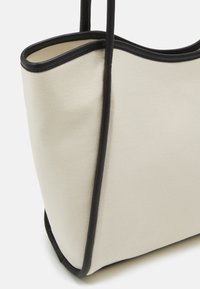 Torba płócienna w kolorze kremowym z czarnym wykończeniem, o miękkiej teksturze i otwartym topie, z wyraźnie wyprofilowanym kształtem i usztywnionym dnem.