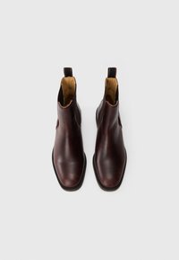 Une paire de bottes Chelsea en cuir brun foncé avec des panneaux élastiques noirs sur les côtés et des languettes de tirage, présentée de dessus sur un fond blanc.