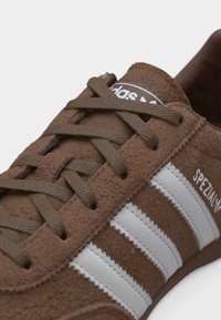 Καφέ σουέντ αθλητικό παπούτσι Adidas Spezial με λευκές ρίγες και καφέ κορδόνια που ταιριάζουν, σε λευκό φόντο.