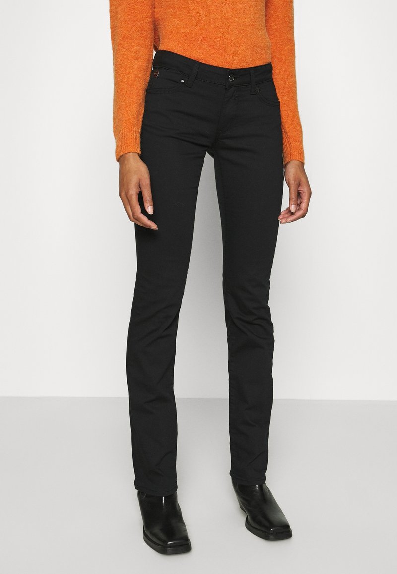 Vaqueros negros ajustados con cinco bolsillos, lazos para cinturón y cierre de botón. Combinados con un suéter naranja texturizado y botas negras.