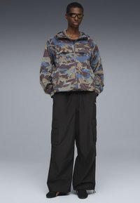 Giacca con zip a motivo camouflage, dotata di cappuccio, caratterizzata da tonalità di marrone, blu e verde, abbinata a pantaloni cargo neri e scarpe nere.
