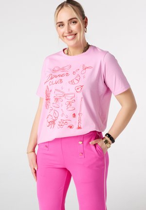 Vrouw glimlachend, draagt een lichtroze "Dinner Club" T-shirt met illustraties van eten en drinken, gecombineerd met felroze broek en gouden sieraden.