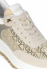 Beige sneakers met suède en meshpanelen, decoratieve metalen studs en een dikke witte zool. Accenten omvatten een zilveren logo op de tong.