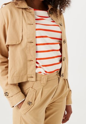 Mujer con chaqueta beige abotonada y pantalones a juego, con una camisa blanca con rayas naranjas metida, mano en el bolsillo.