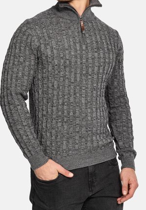 Hombre con suéter de punto gris texturizado con un cuello corto con cremallera y pantalones negros, con una mano en el bolsillo.