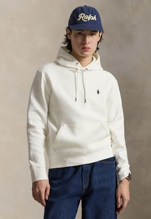 Ung mand iført en hvid hættetrøje med et lille sort logo, blå jeans, en mørkeblå kasket med teksten "Ralph" og et armbåndsur på en neutral baggrund.