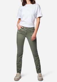 Pantalones de algodón verdes con corte ajustado, bolsillos frontales y cierre de botón, combinados con una camiseta de cuello redondo blanca y zapatillas ligeras.