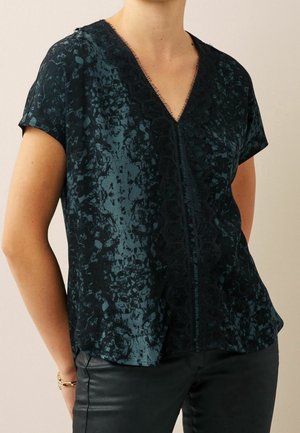 Femme portant un chemisier à manches courtes bleu sarcelle foncé à motifs avec un col en V orné de dentelle noire, associé à un pantalon noir et un bracelet au poignet.