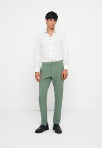 White button-up shirt with blue accents, paired with tailored green pants and black shoes, on a neutral background.