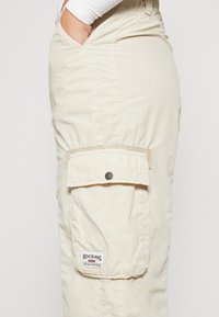 Beige cargo bukser med en sidelomme med trykknapp og et stoffmerke. Laget av et teksturert, lett materiale.
