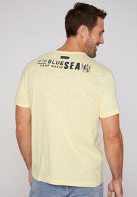 Camiseta amarilla de manga corta con un acabado texturizado y un estampado gráfico en la parte posterior. Etiqueta cerca del cuello, detallado en las costuras laterales.