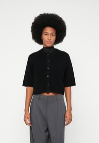 Black cropped cardigan with short sleeves, round collar, and button front, made from soft material. Paired with gray high-waisted trousers.