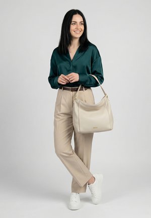 Nainen, jolla on mustat hiukset, pukeutunut tummaan vihreään puseroon ja beigenvärisiin housuihin, kantaa suurta beigeä käsilaukkua ja pukeutunut valkoisiin tennareihin.