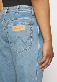 Person som bär ljusblå Wrangler-jeans med en synlig bakficka och lädermärke, hand avslappnat vid sin sida.