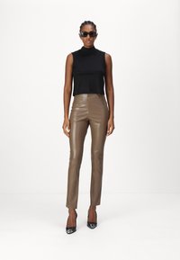 Top senza maniche nero con collo alto finto, pantaloni in finta pelle marrone e décolleté neri a punta, su uno sfondo bianco semplice.
