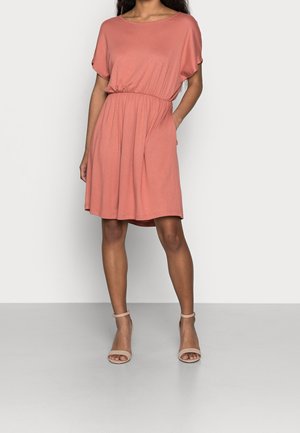 Femme portant une robe corail à manches courtes avec taille élastique et des sandales beige à bride cheville, debout devant un fond uni.