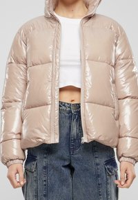 Wypchana, błyszcząca, beżowa kurtka puchowa z kapturem, krótka, z zamkiem z przodu i elastycznymi mankietami, zestawiona z dżinsowymi spodniami o wysokim stanie.