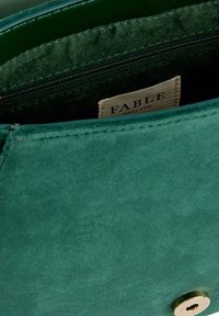Grön mocka handväska med ett foder som har en dragkedjeficka och en sydd etikett som läser "FABLE ENGLAND." Har en slät textur och cirkulär hårdvarudetalj.